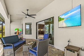 A covered outdoor seating area with wicker furniture, a ceiling fan, and a television displaying a beach scene. - Dog-Friendly Waterfront Escape with Heated Pool & Outdoor Grill (Cape Coral)