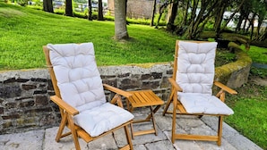 Two white cushioned chairs with wooden frames and a small wooden table, placed on a stone-paved area with a grassy background and trees. - Pylemoor Cottage (Tiverton)