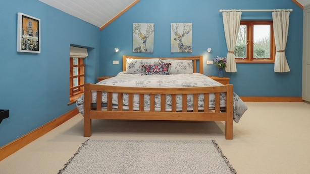 A bedroom with a wooden bed, blue walls, a window with curtains, and two wall-mounted paintings. - Pylemoor Cottage (Tiverton)