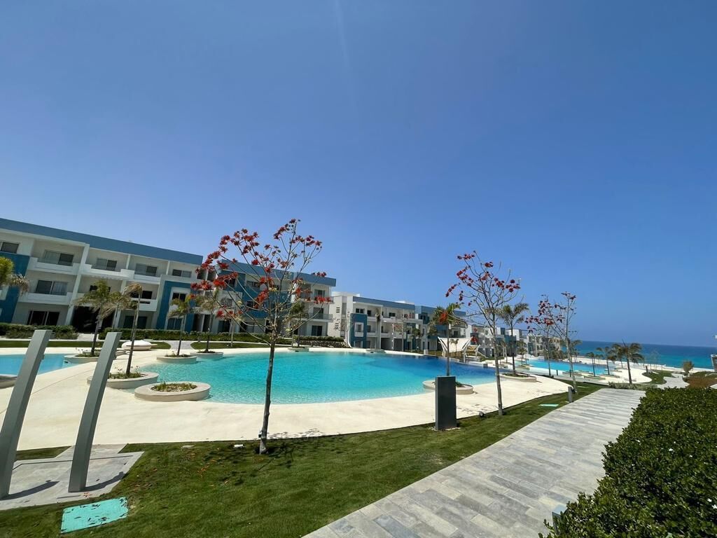 Area piscina di un hotel con cielo azzurro, una grande vasca e un edificio con numerosi balconi.
