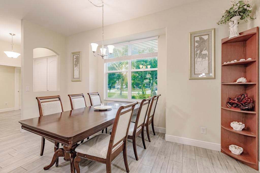 A dining room with a large wooden table, six chairs, a chandelier, a window with blinds, and a wooden shelving unit with decorative items.