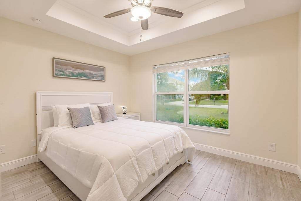A bedroom with a bed, a ceiling fan, a window with a view of greenery, and a painting on the wall.