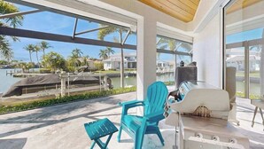 A modern outdoor patio with a grill, two turquoise chairs, and a view of a marina with boats and palm trees. - Spacious canal front home with private pool, dock & close to everything (Marco Island)