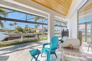 A modern outdoor patio with a grill, two turquoise chairs, and a view of a marina with boats and palm trees. - Spacious canal front home with private pool, dock & close to everything (Marco Island)