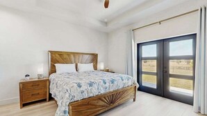 Un dormitorio con cama, mesita de noche, ventilador de techo y un amplio ventanal con cortinas.
