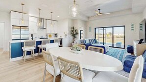 Una cocina moderna con armarios blancos, un comedor con una mesa redonda y una sala de estar con un sofá y un ventanal grande.