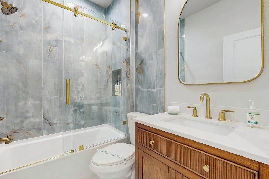 A bathroom with a glass-enclosed shower, a white toilet, a wooden vanity with a gold faucet, and a large mirror.