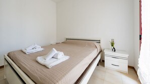 A neatly made bed with a beige bedspread and two rolled towels, a white bedside table with a vase of flowers, and a white nightstand with drawers. - GuestHost - Alba Cozy Flat near the Sea W/Parking (Alba Adriatica)