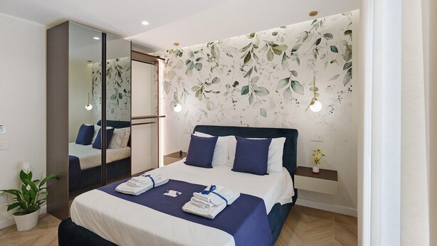Une chambre d’hôtel avec un lit, un miroir, une table de chevet, une plante en pot et un mur orné d’un motif de feuilles.