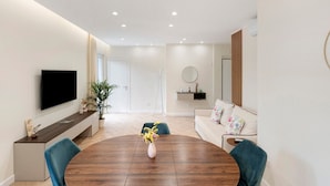Un salon moderne avec une table à manger ronde en bois, un téléviseur à écran plat fixé au mur, un canapé agrémenté de coussins à motifs floraux et une plante en pot.