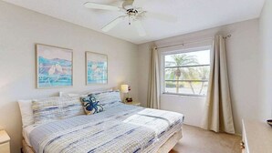 Een slaapkamer met een bed, een plafondventilator, twee ingelijste foto's, een raam met gordijnen en een nachtkastje met een lamp.