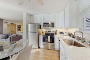 A modern kitchen with white cabinets, stainless steel appliances, a glass dining table, and a view of the living area. - Gorgeous condo with modern upgrades, Gulfviews & shared pool (Bonita Springs)