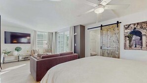 Una habitación de hotel moderna con una cama grande, un área de descanso con sofá y sillas, un ventilador de techo y un ventanal amplio.