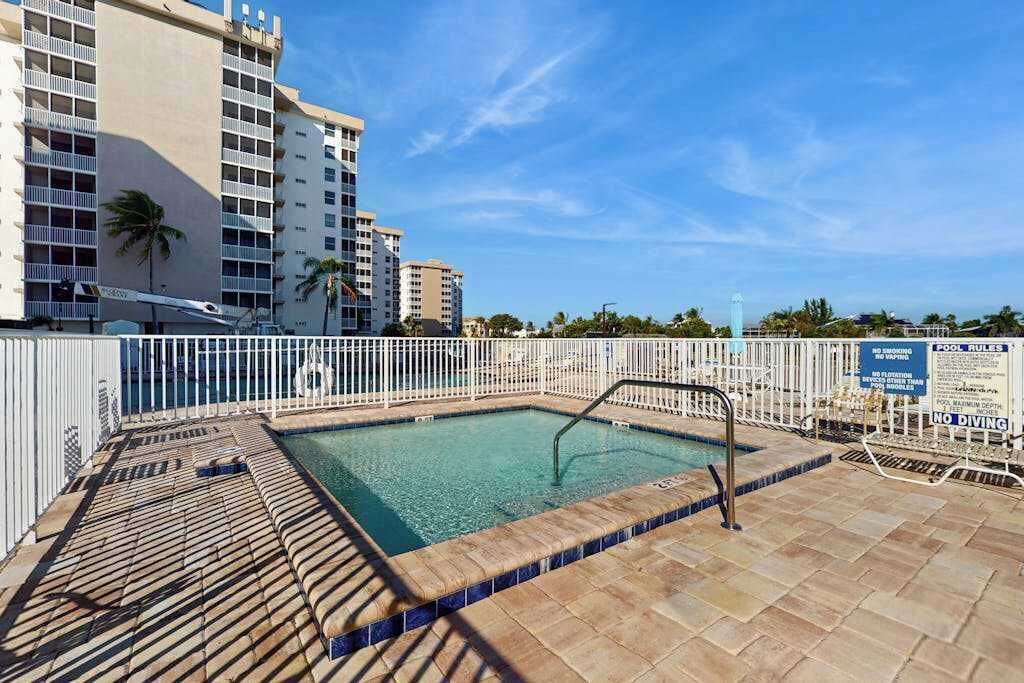 A swimming pool with a tiled deck, surrounded by a white fence, and adjacent to a multi-story building.
