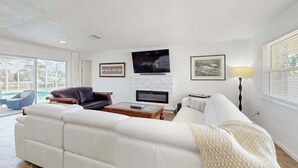 A living room with a white sofa, a black leather chair, a fireplace, a TV, and framed pictures on the wall. - Snowbird Retreat – Canal Home w/Pool, Game Room & Pet Friendly (Punta Gorda)