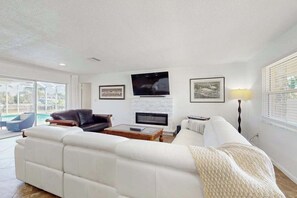 A living room with a white sofa, a black leather chair, a fireplace, a TV, and framed pictures on the wall. - Snowbird Retreat – Canal Home w/Pool, Game Room & Pet Friendly (Punta Gorda)