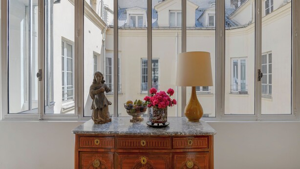 A room with a large window, a wooden cabinet with drawers, a statue, a vase with flowers, and a lamp. - Paris Opera Prestige residence (Paris)