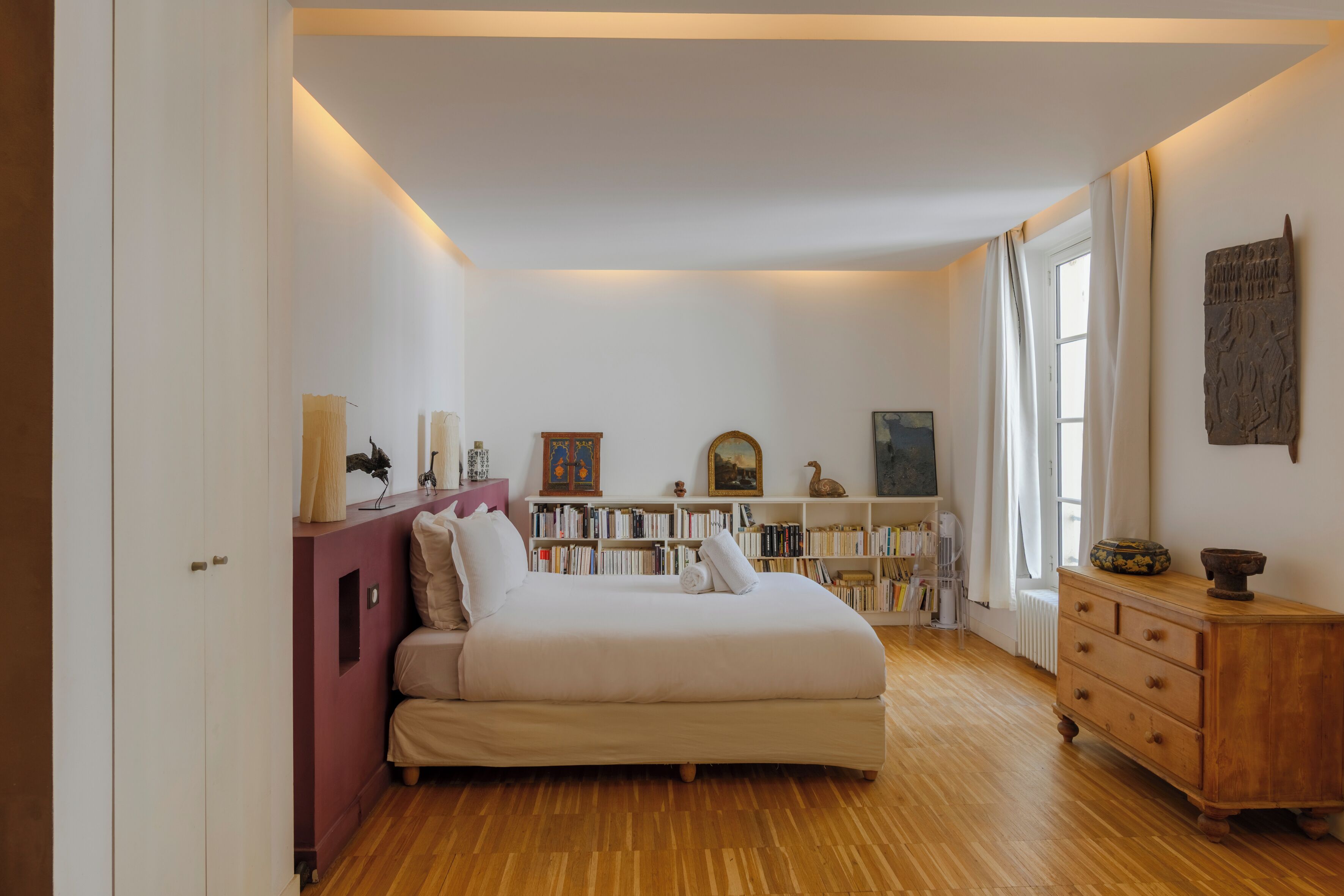 Een slaapkamer met een bed, een houten dressoir, een boekenkast en een raam met gordijnen.
