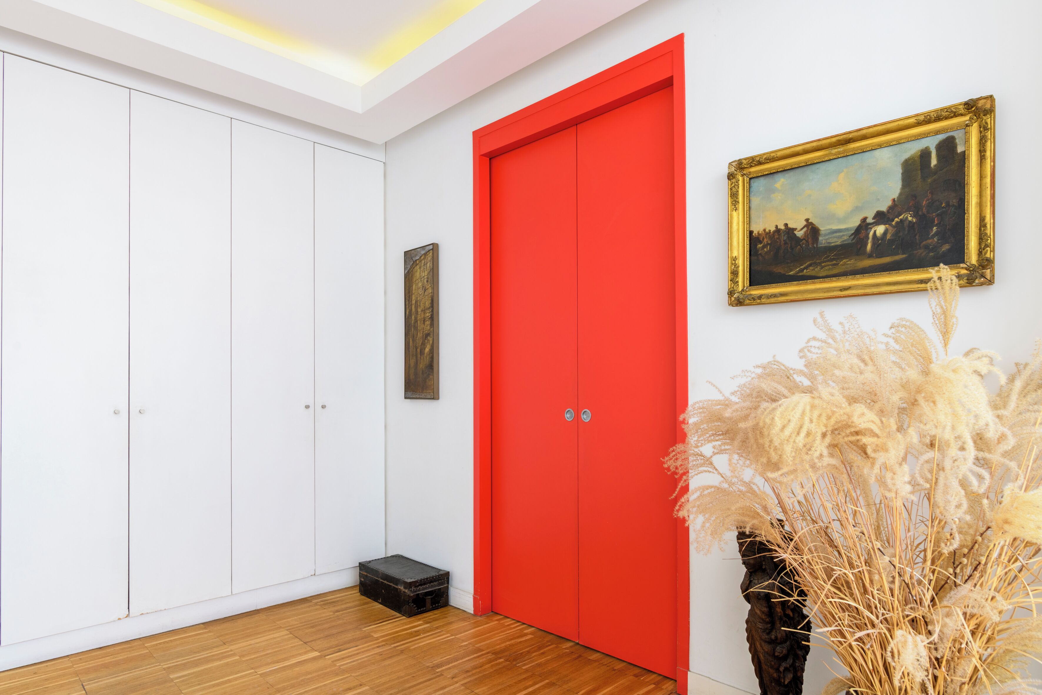 A room with a white wardrobe, a red door, a framed painting, and a decorative plant.