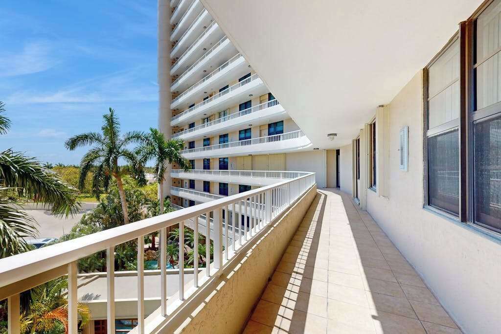 Balcón de un hotel de varios pisos con suelo de baldosas, barandilla blanca y vistas a palmeras.