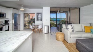 A modern kitchen with a marble countertop, a dining area with chairs, a sofa with cushions, and a view of the ocean through large windows. - Amazing oceanfront condo with pool, hot tub, tennis, & stunning balcony views (Marco Island)