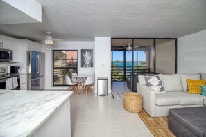 A modern kitchen with a marble countertop, a dining area with chairs, a sofa with cushions, and a view of the ocean through large windows. - Amazing oceanfront condo with pool, hot tub, tennis, & stunning balcony views (Marco Island)