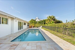 Une piscine avec une terrasse carrelée, un bâtiment blanc doté de grandes fenêtres et un espace clos agrémenté de verdure.