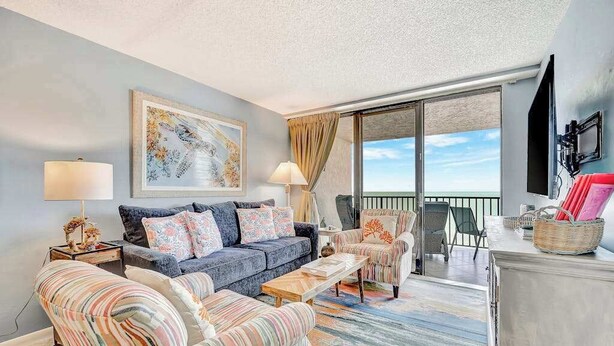 Ein Wohnzimmer mit einer Couch, Sesseln, einem Couchtisch, einem Fernseher und einem Balkon mit Blick.