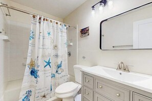 Un bany amb un vàter blanc, un lavabo amb mirall i una cortina de bany amb motius de estrelles de mar i criatures marines.