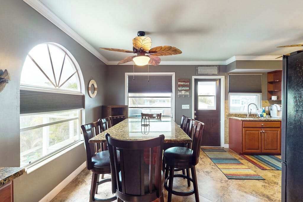 Una cocina con isla central, sillas de madera oscura, ventilador de techo y una ventana con persianas.