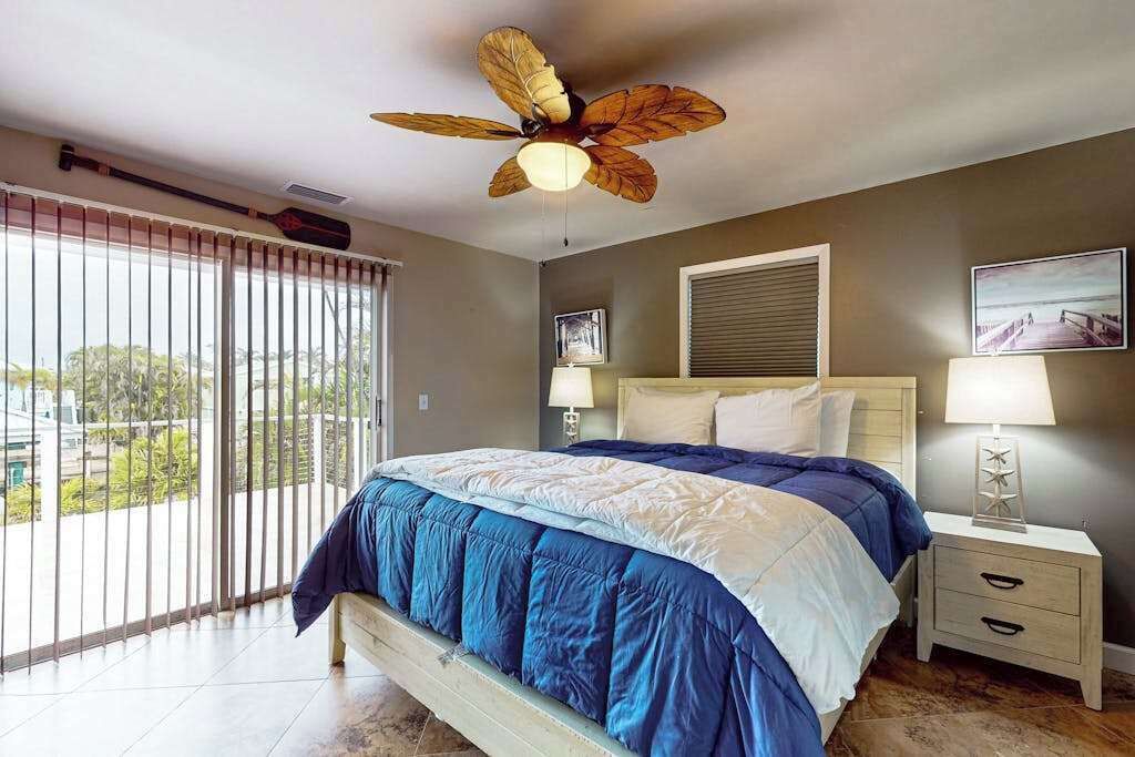 A bedroom with a bed, a ceiling fan, a nightstand, a window with blinds, and a view of the outside.