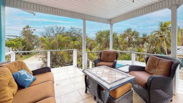 A covered outdoor seating area with wicker furniture and a glass-top table, overlooking a tropical landscape. - Spacious home with heated pool, lots of outdoor entertainment, and WIFI (Fort Myers Beach)