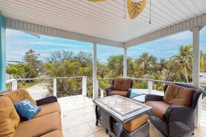 A covered outdoor seating area with wicker furniture and a glass-top table, overlooking a tropical landscape.