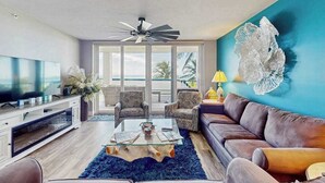 A modern living room with a television, leather sofa, patterned chairs, a glass coffee table, and a decorative wall piece. - Beachfront condo with huge lanai, sunset gulf views, pool, hot tub, tennis & gym (Marco Island)
