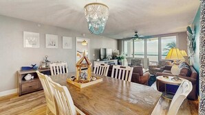A dining area with a wooden table, chairs, a chandelier, and a view of the ocean through large windows. - Beachfront condo with huge lanai, sunset gulf views, pool, hot tub, tennis & gym (Marco Island)