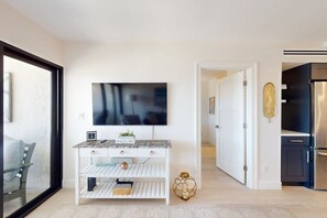 A modern living room with a flat-screen TV, a white console table, a sofa, and a kitchen area with a refrigerator. - Remodeled condo in a waterfront complex with pools, hot tubs, & tennis (Marco Island)