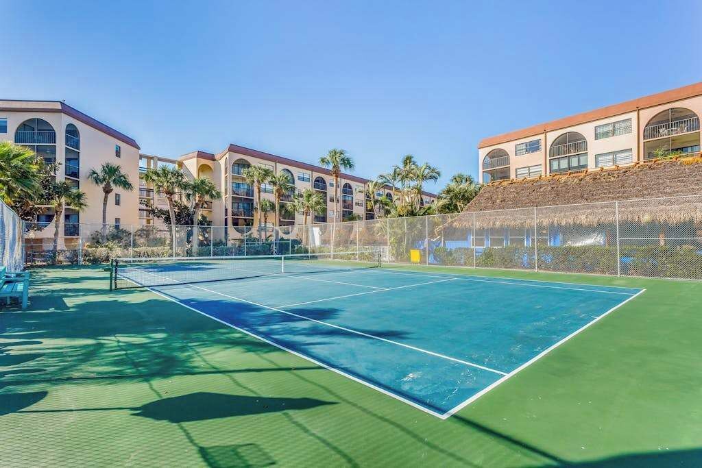 A tennis court with a net, surrounded by a fence, and adjacent to a building with multiple balconies and palm trees.