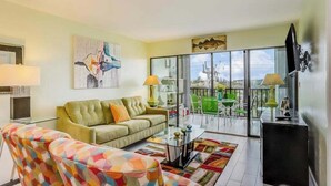 Un salon avec un canapé, un fauteuil et une table en verre. Il y a un balcon avec des chaises et un téléviseur.