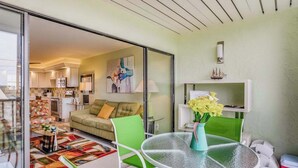 Un salon moderne avec une cloison en verre, un canapé vert, une table ronde en verre surmontée d’un vase, et une cuisine en arrière-plan.