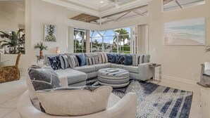 A modern living room with a sectional sofa, a round ottoman, and a large window offering a view of palm trees. - Waterfront home with saltwater pool, pool spa, central AC, W/D, & office space (Marco Island)