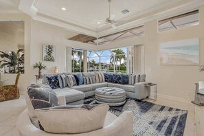 A modern living room with a sectional sofa, a round ottoman, and a large window offering a view of palm trees. - Waterfront home with saltwater pool, pool spa, central AC, W/D, & office space (Marco Island)
