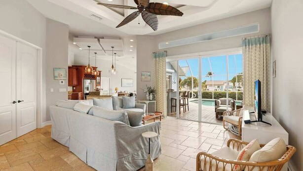 A spacious living area with a sofa, a dining table, and a TV. There is a ceiling fan, a kitchen in the background, and a view of the outdoors. - Large corner lot home with canal views & screened in pool - close to the beach (Marco Island)