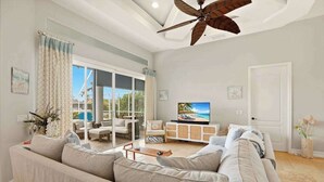 A spacious living room with a large sofa, a television, and a ceiling fan. - Large corner lot home with canal views & screened in pool - close to the beach (Marco Island)