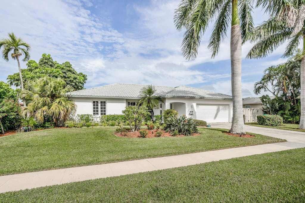 A single-story house with a white exterior, a garage, and a well-maintained lawn with palm trees.