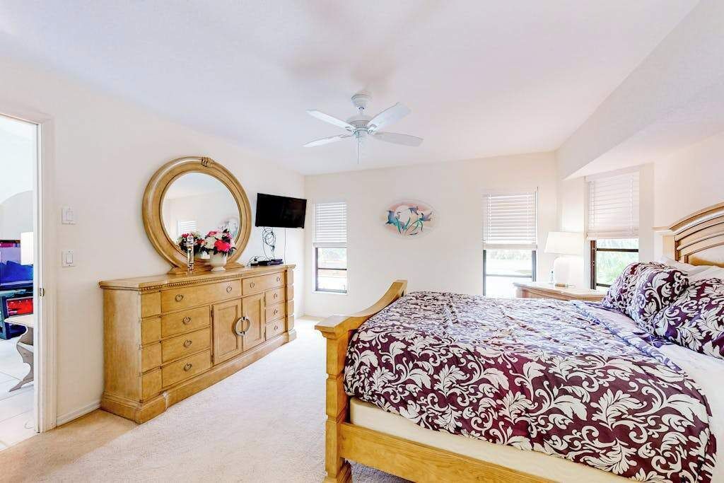 A bedroom with a bed, dresser, mirror, ceiling fan, and windows.