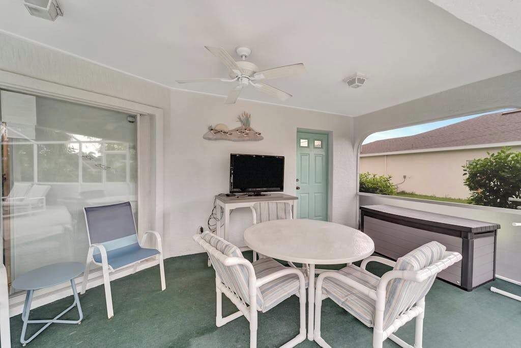 A balcony with a white table, four chairs, a ceiling fan, a TV, and a door.