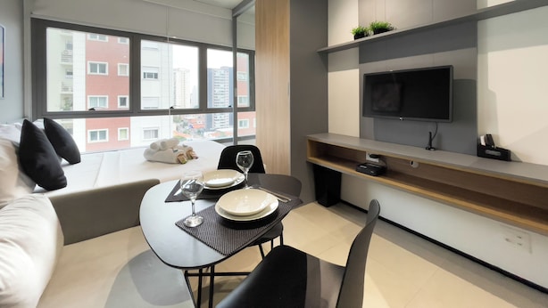 Moderni hotellihuone, jossa on ruokailutila, seinälle asennettu taulutelevisio ja suuri ikkuna, josta avautuu näkymä kaupunkiin.