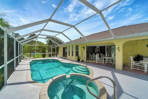 A covered outdoor swimming pool with a glass ceiling, a patio area with chairs, and a yellow building with a tiled roof. - Relax & Recharge in This Spacious Port Charlotte Retreat with Pool & More! (Port Charlotte)