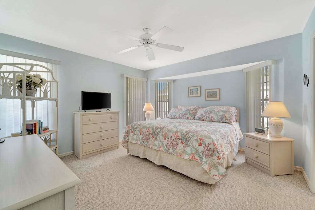 A bedroom with a bed, a desk, a television, a ceiling fan, and a window with a view.