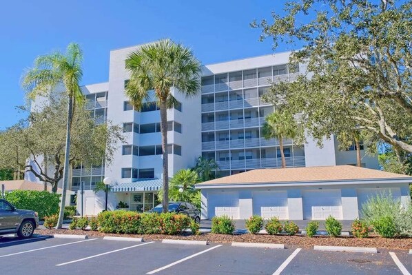 A multi-story hotel with a modern design, featuring balconies, a parking lot, and surrounding greenery. - 3rd-floor snowbird-friendly condo with enclosed balcony, outdoor pool, W/D, AC (Naples)